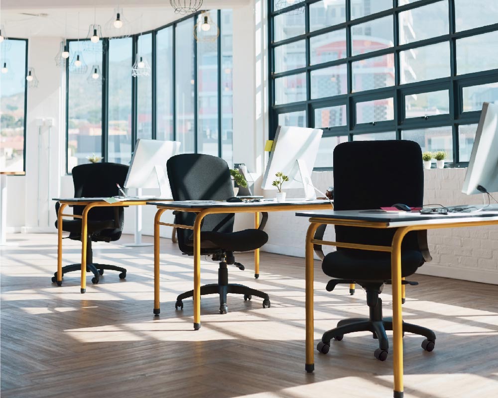 Modern office space with large windows, featuring desks and black office chairs with caster wheels arranged neatly in rows.